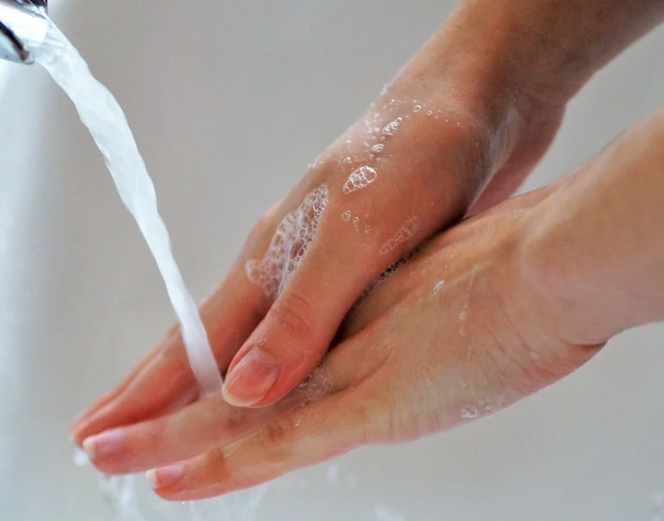 Descrição da imagem: Mulher lavando as mãos, em uma pia branca, com água caindo.